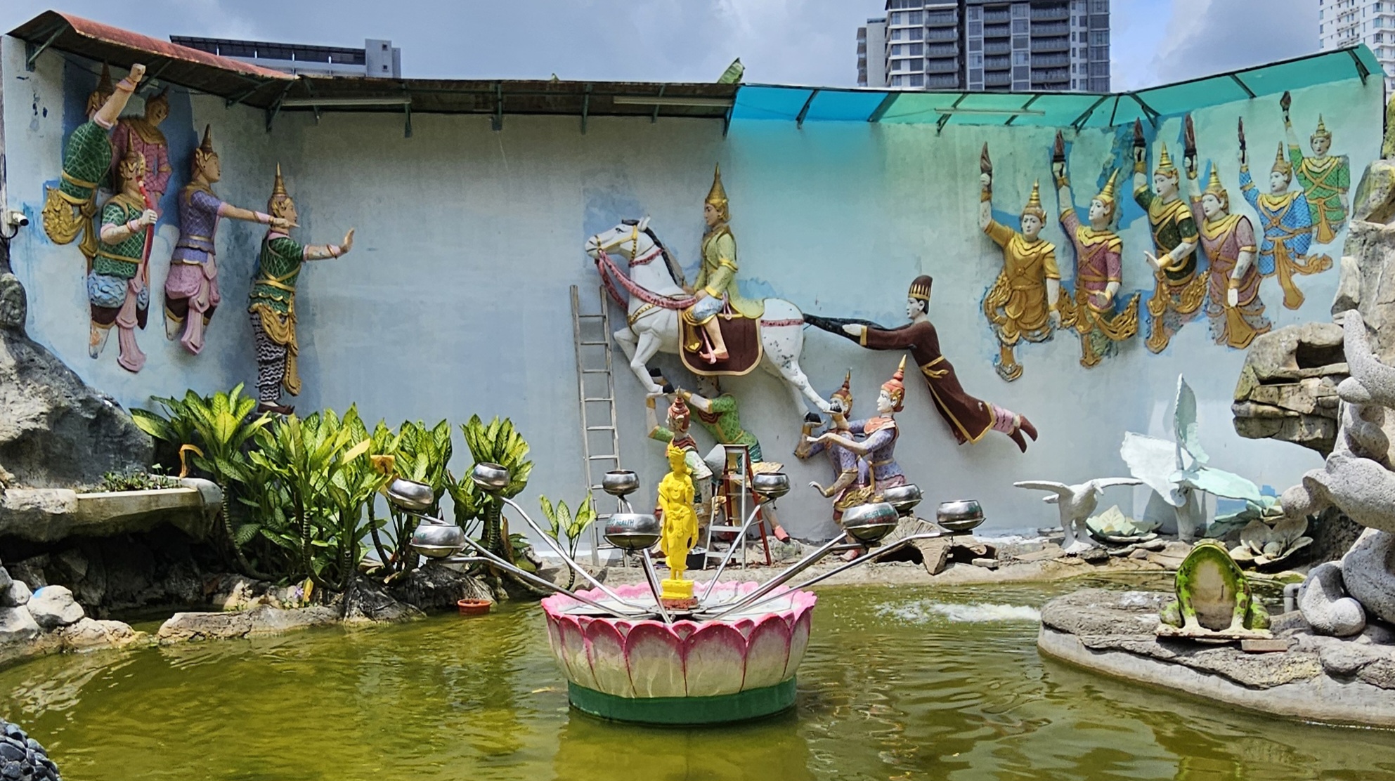 A scenic area featuring three-dimensional wall murals of mythical figures, a central pond with a lotus-shaped pedestal and a golden statue, surrounded by lush greenery and additional decorative elements.