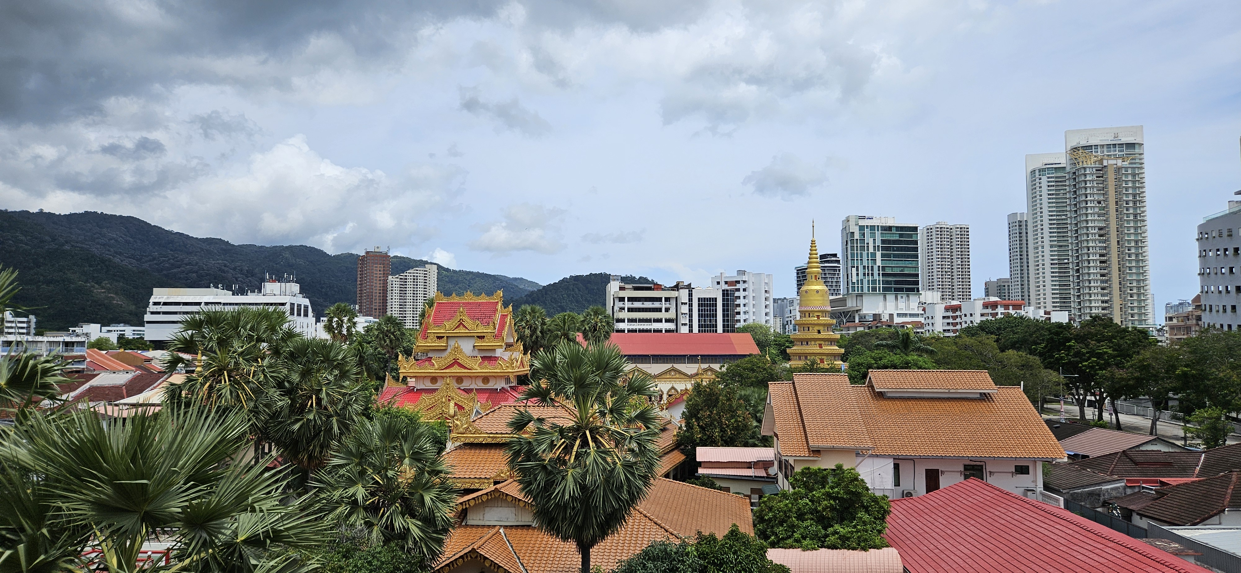 A panoramic view of a cityscape featuring traditional Asian architecture with ornate rooftops, surrounded by modern buildings and lush greenery under a cloudy sky.