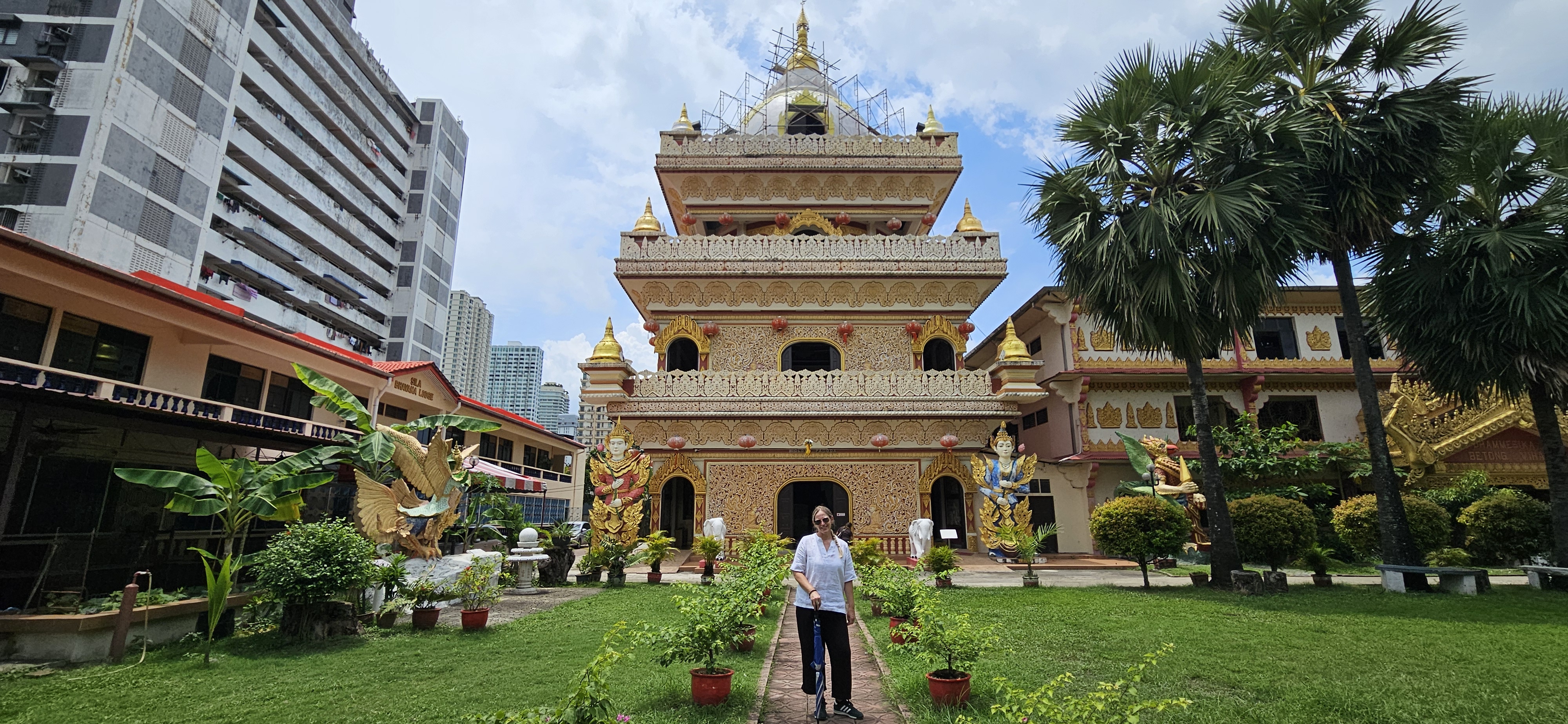 A woman standing in front of an ornate temple with intricate designs, surrounded by greenery and palm trees, with modern buildings in the background.