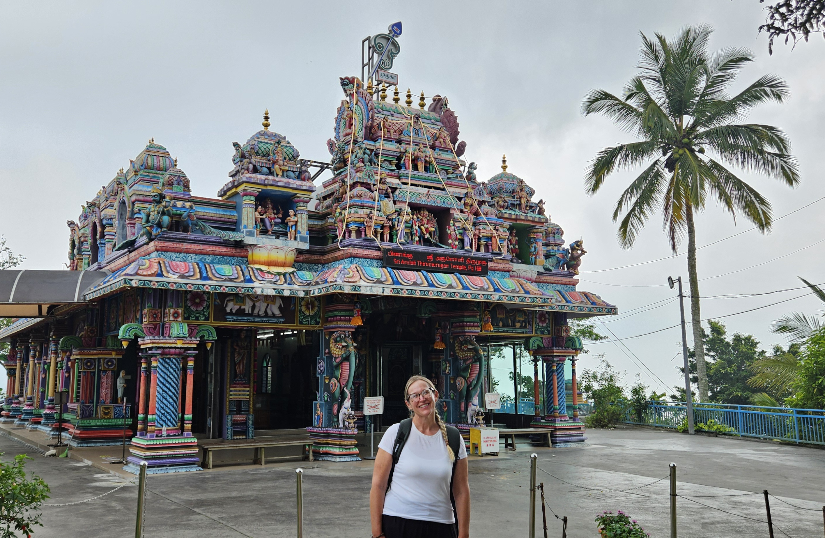 A person standing in front of a colorful Hindu temple adorned with intricate sculptures and designs, set against a cloudy sky with palm trees nearby.