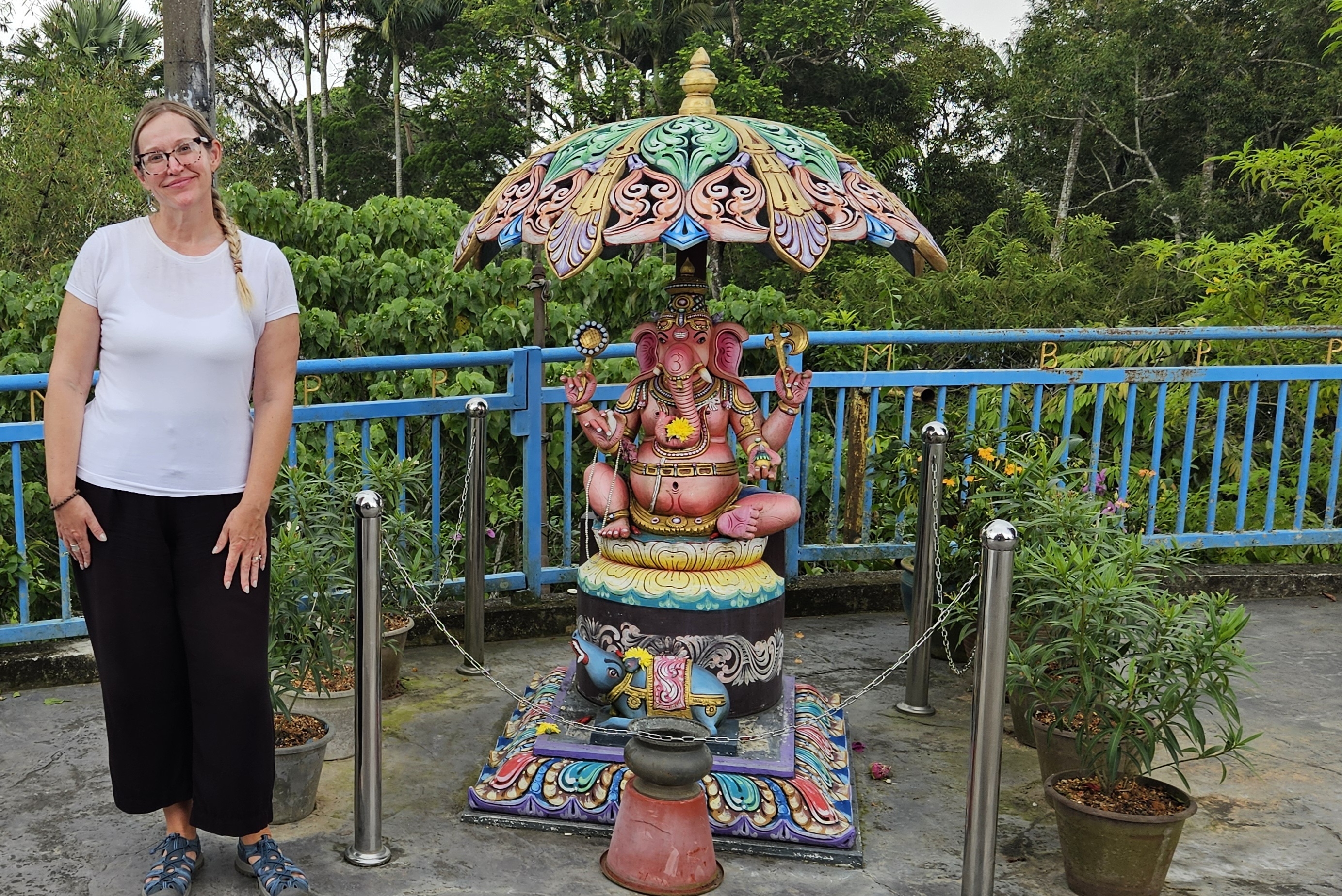 A woman stands beside a colorful statue of Lord Ganesha, adorned with decorations and an umbrella, in a lush green outdoor setting.