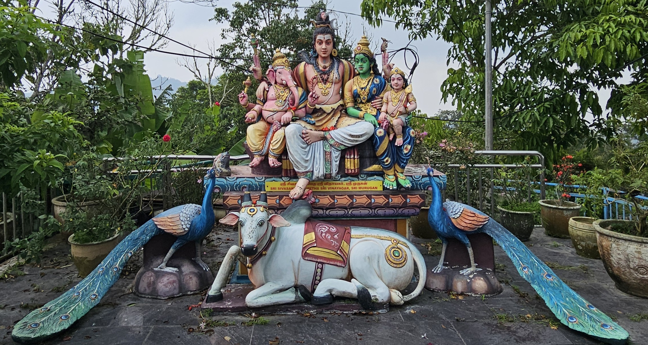 A colorful sculpture depicting a deity surrounded by various figures, including a cow and peacocks, set in a lush garden with greenery and flowers in the background.