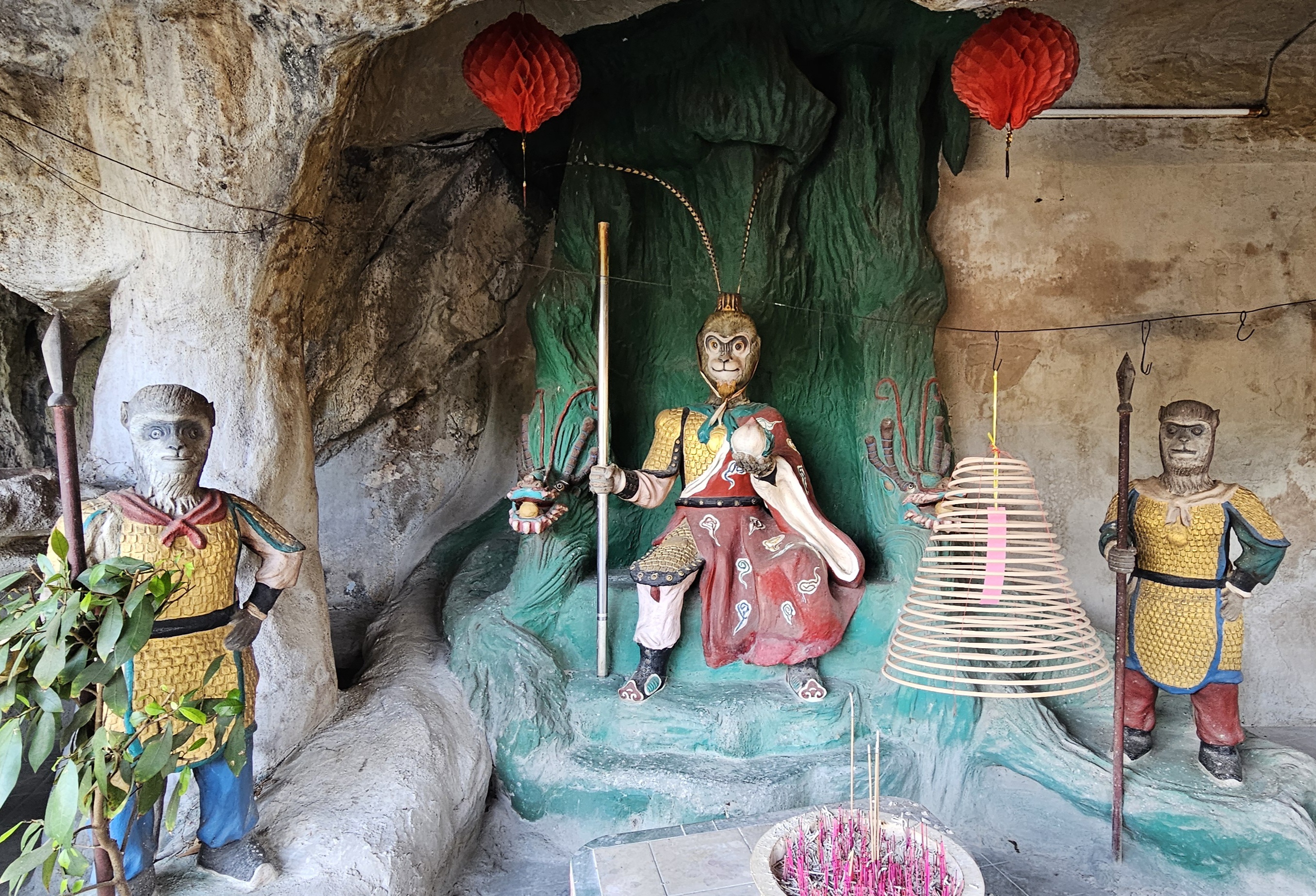 A scene featuring three colorful statues in a cave setting. The central statue is of a monkey-like figure dressed in traditional attire, holding a staff. The left and right statues are also monkey figures, each wielding a spear. There is a green rock wall behind them, and red lanterns and incense stick holders are present in the surroundings.