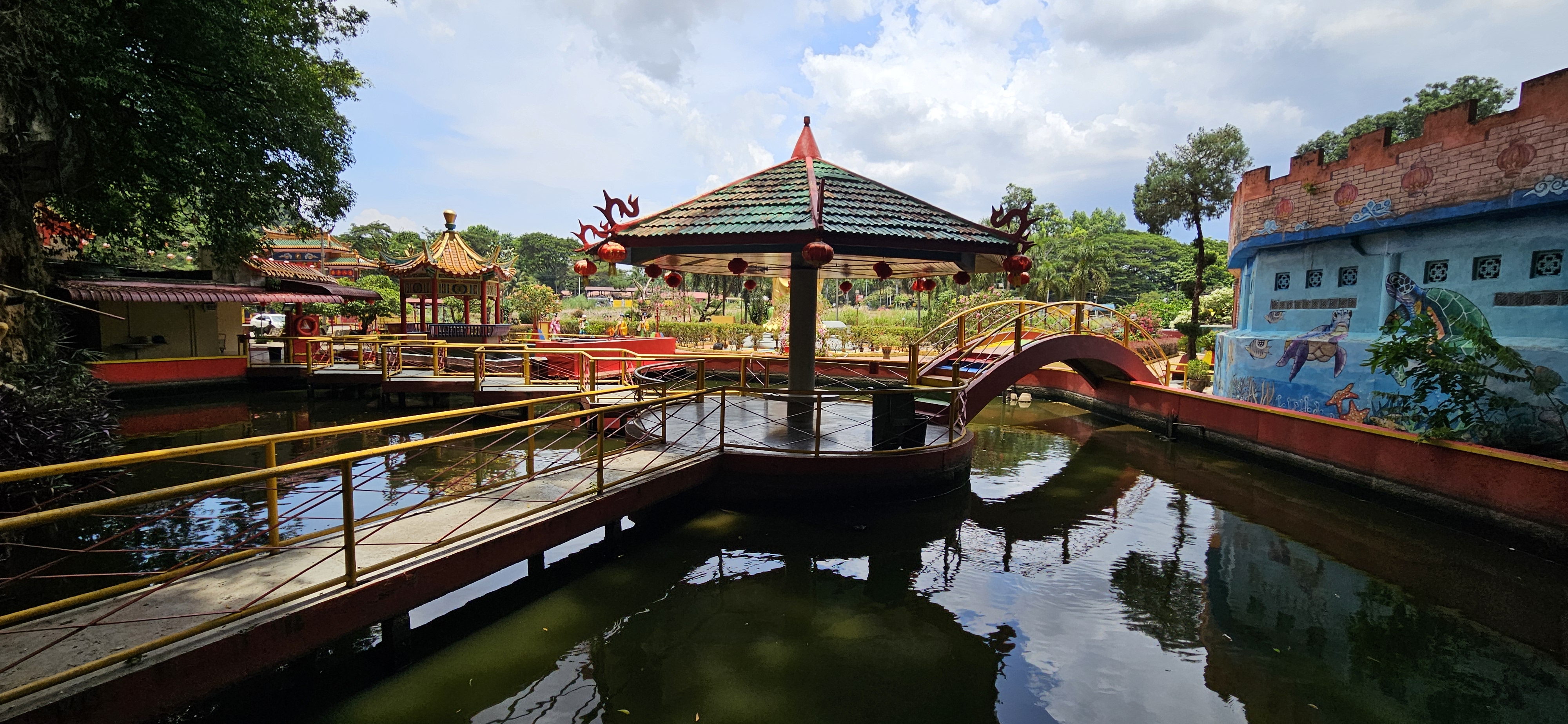A scenic view of a tranquil park featuring a gazebo surrounded by water, decorative bridges, and lush greenery under a partly cloudy sky.