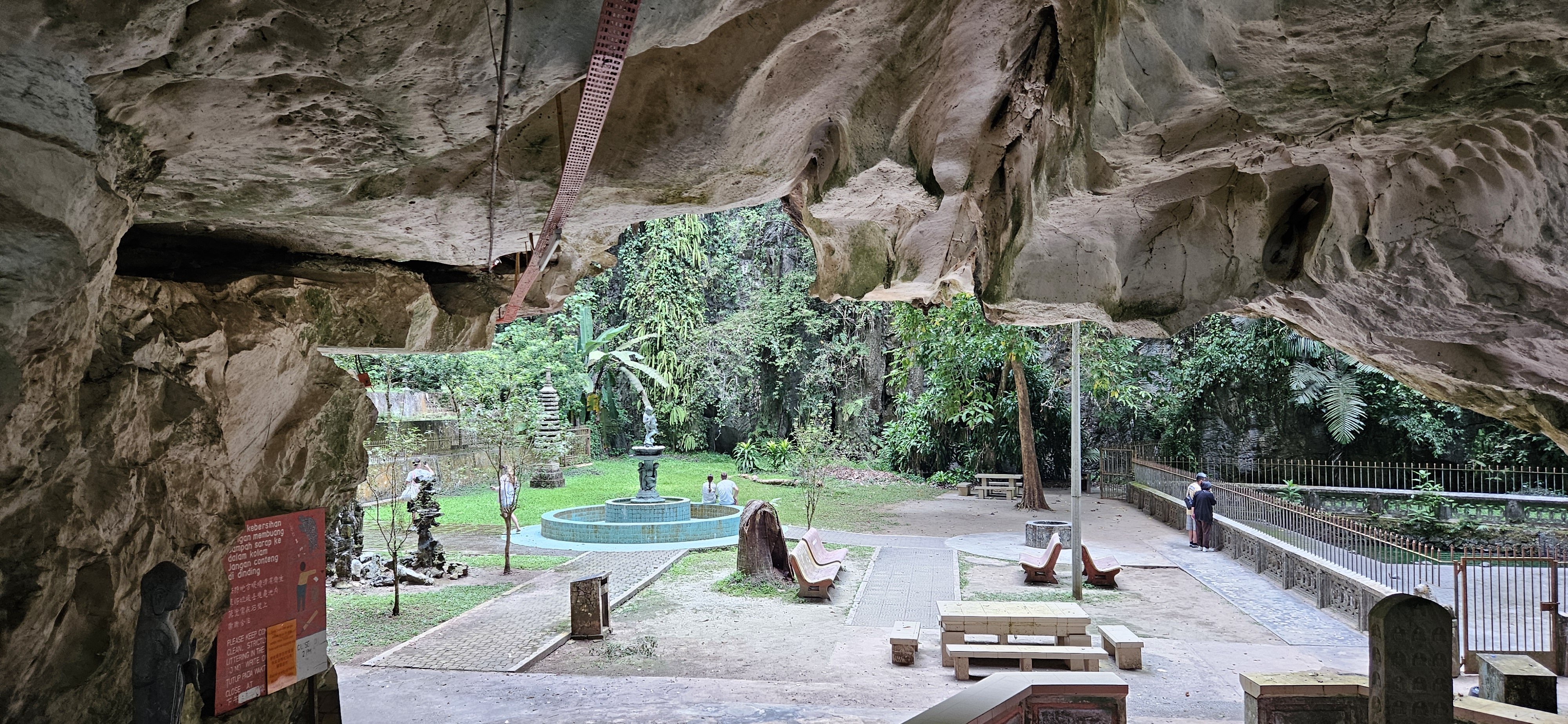 View from inside a cave entrance looking out onto a lush green garden with a stone fountain, seating areas, and sculptures.