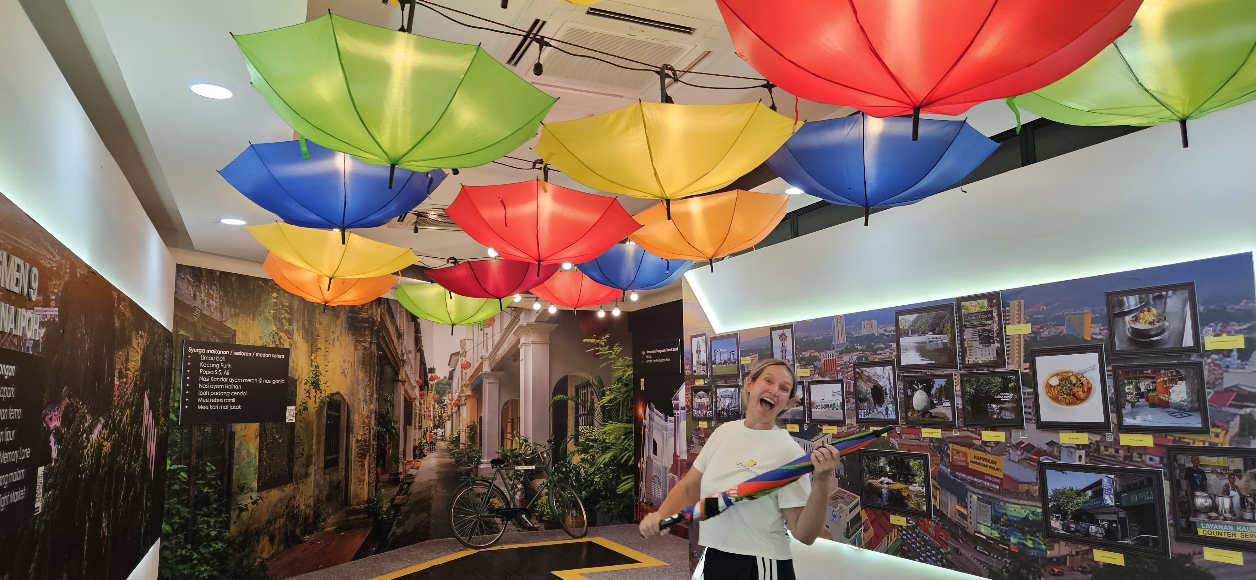 A woman posing happily in a colorful indoor space decorated with hanging umbrellas. The background features a mural of a street lined with vibrant buildings and a bicycle, along with framed photos of local attractions and food.