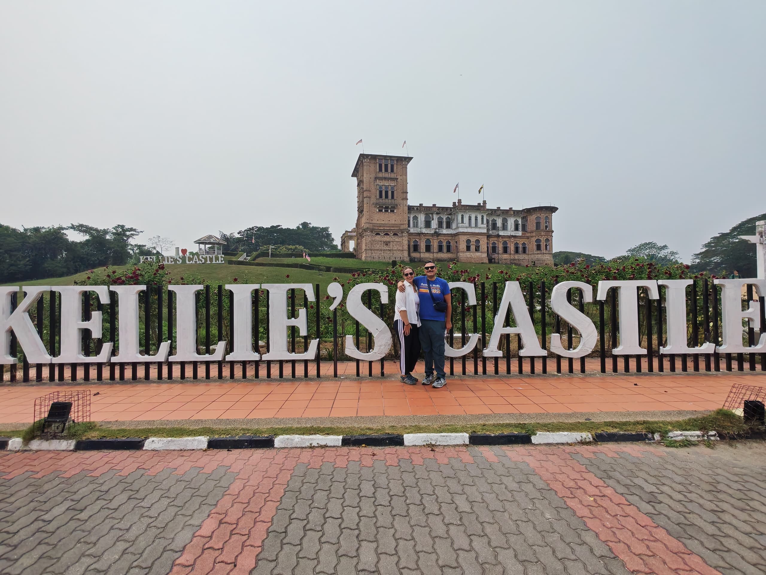 A couple stands in front of the sign for Kellie's Castle, with the castle visible in the background. The sign features large, bold letters spelling 'KELLIE'S CASTLE'.
