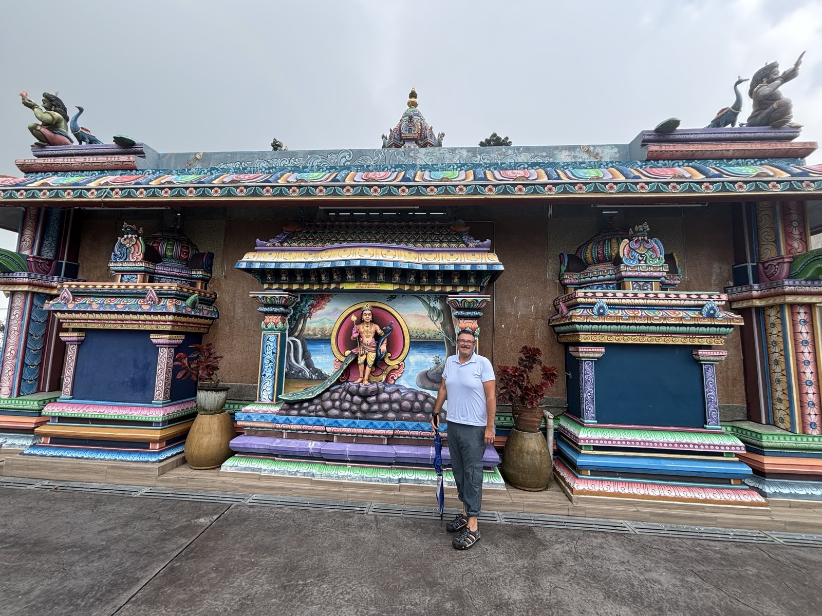 A man standing in front of a colorful temple facade featuring intricate carvings and statues, with vibrant decorations and a mural depicting a deity.