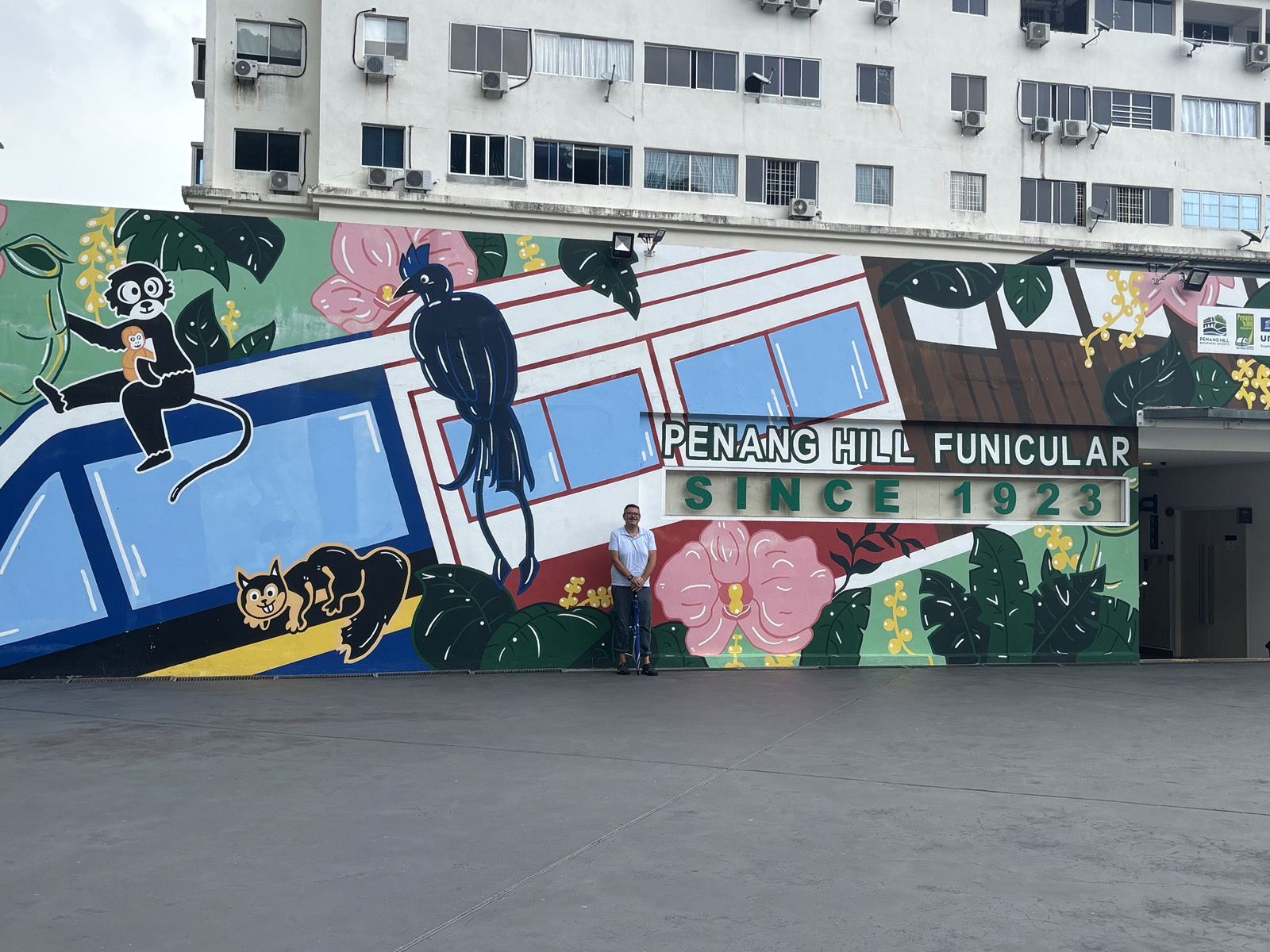 A mural depicting a funicular train with a monkey holding a baby, a black cat, and a bird on a colorful background with flowers, located at the Penang Hill Funicular station. The mural includes the text 'PENANG HILL FUNICULAR SINCE 1923.'