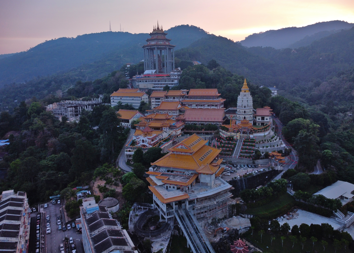 Aerial view of a large, traditional temple complex set in a mountainous landscape at sunset, featuring golden rooftops and surrounded by lush greenery.
