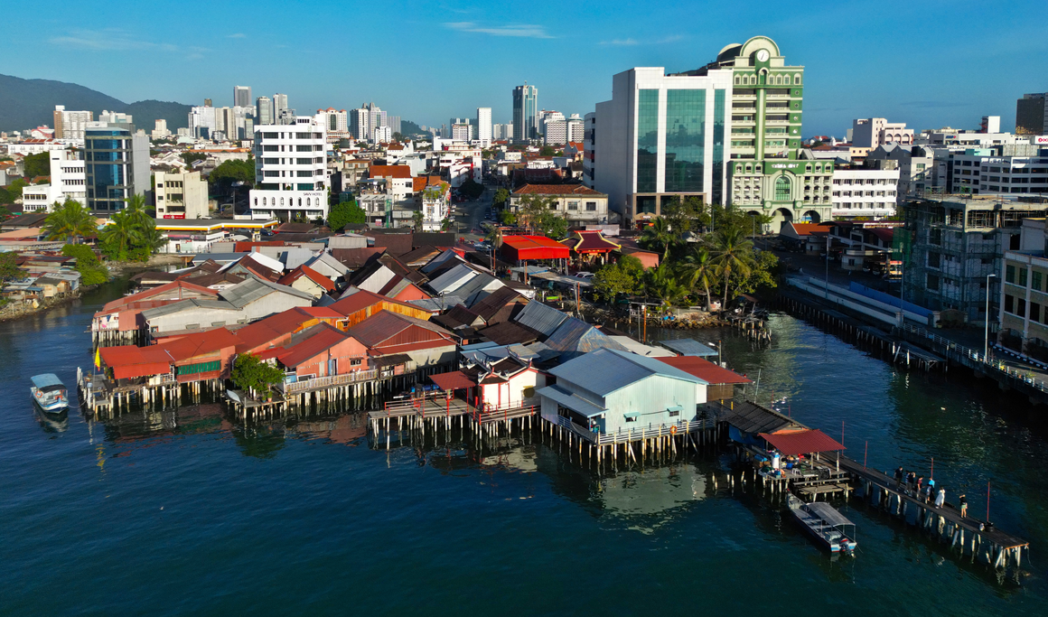 Aerial view of colorful stilt houses along the waterfront, surrounded by modern buildings and skyscrapers in an urban setting.