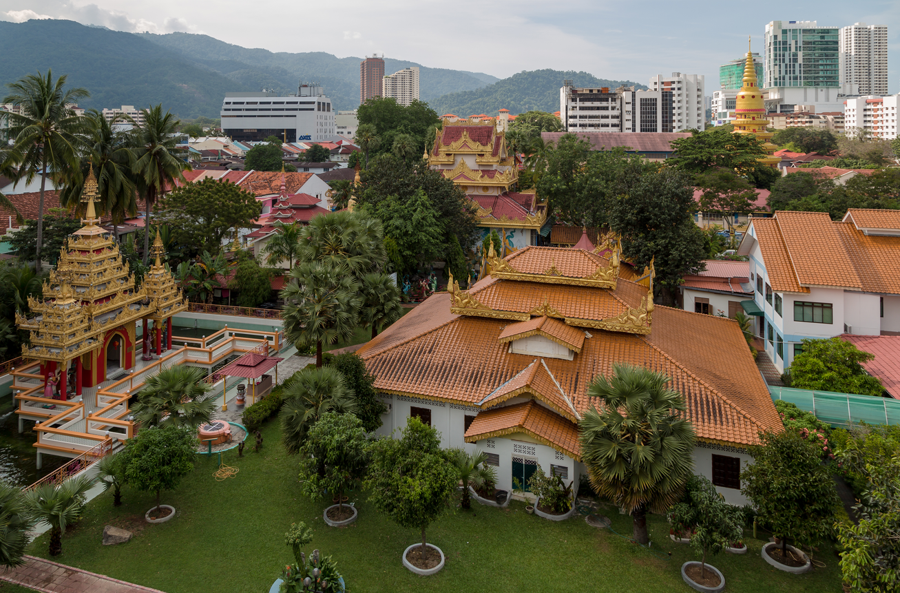 Aerial view of a vibrant neighborhood featuring traditional Asian architecture with ornate golden roofs, surrounded by lush greenery and modern buildings in the background.
